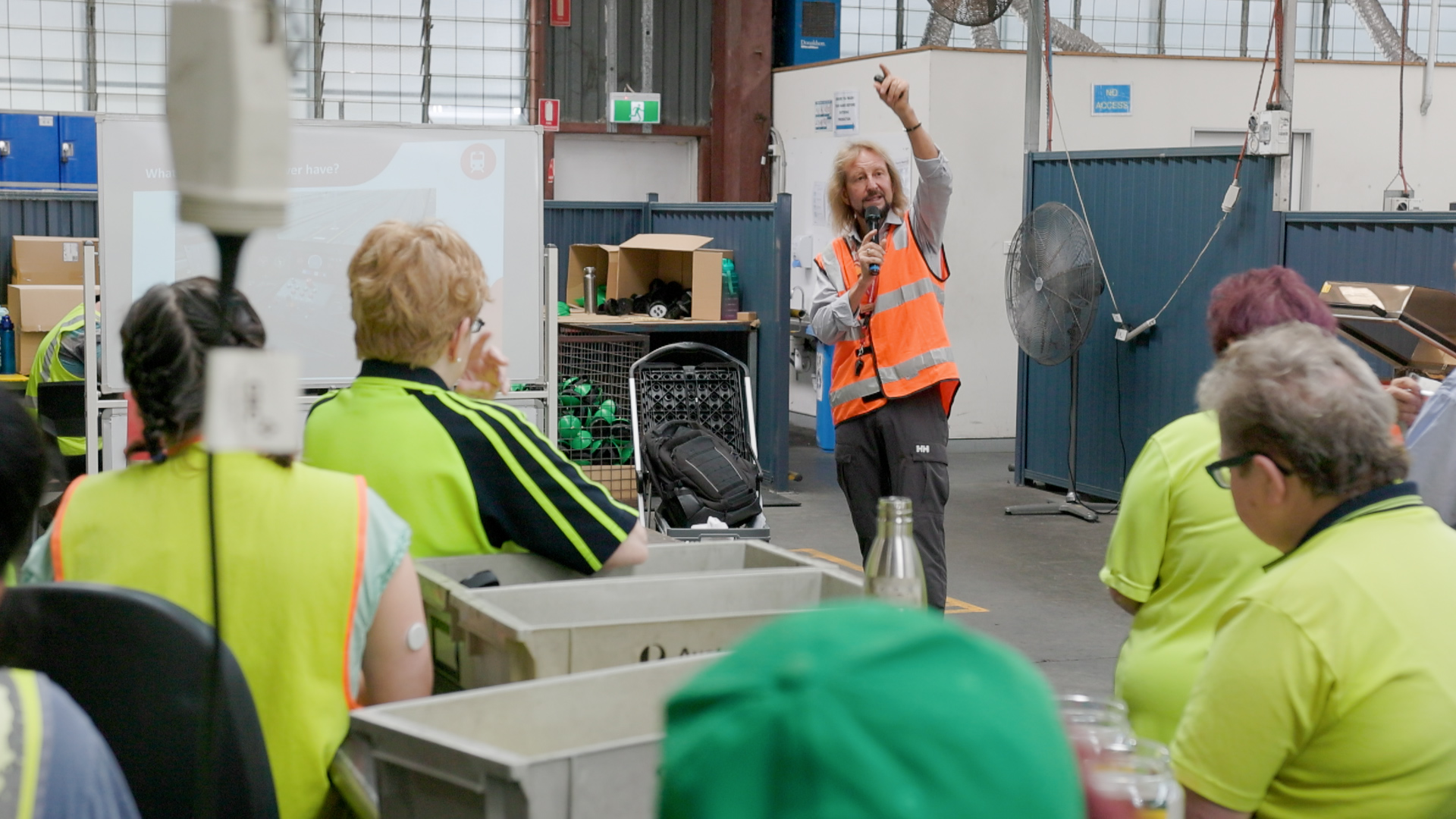 A Community Education Coordinator in a safety vest presenting to a group in a warehouse setting