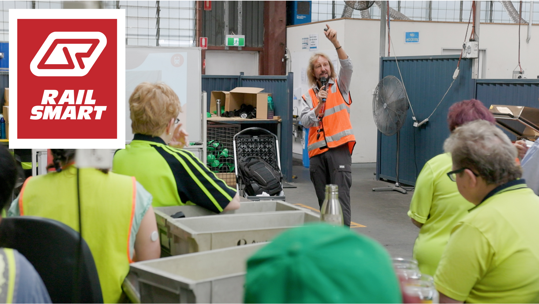 A Community Education Coordinator in a safety vest presenting to a group in a warehouse setting